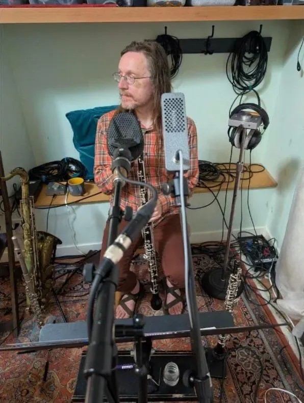 Reed player seated between a ribbon mic and a condenser, saxophone on a stand beside him