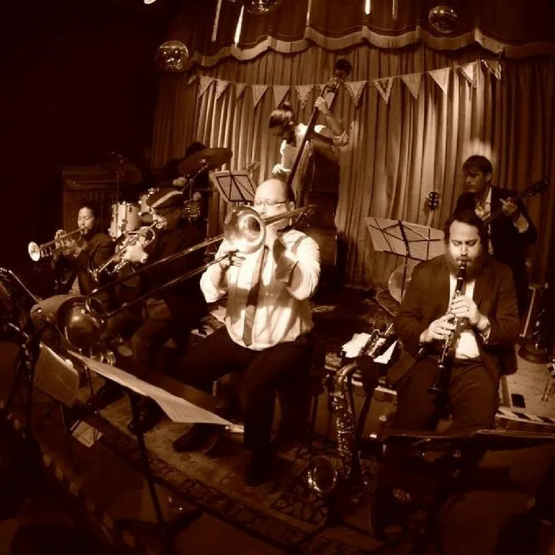 Lucky 7 on stage in sepia, trumpet, trombone and clarinet up front, upright bass and guitar behind, bunting over the curtain