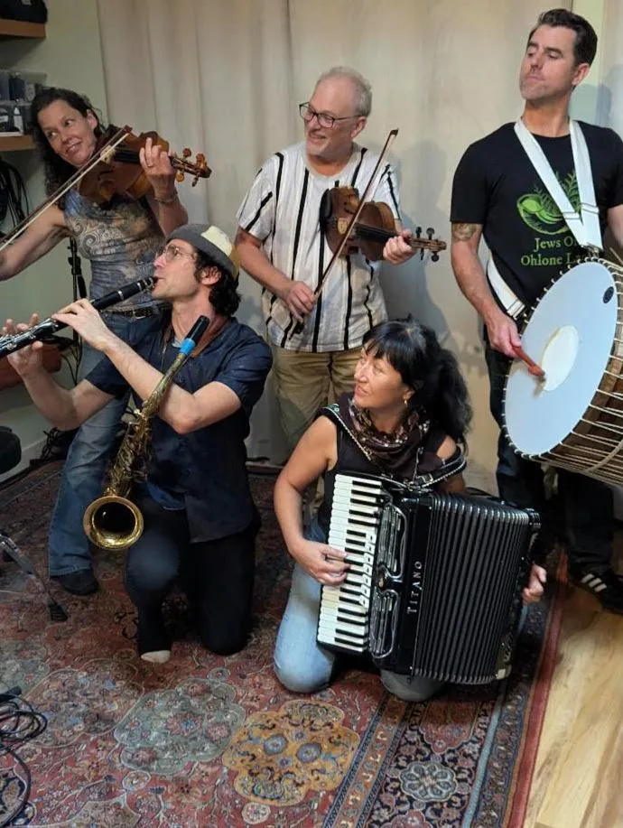 Five musicians with violin, viola, clarinet, accordion and tapan drum gathered on a rug in the live room